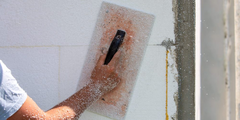 Hand of a worker with a tool for sanding and making smooth and even the surface of a house wall insulated with styrofoam insulation sheets.
